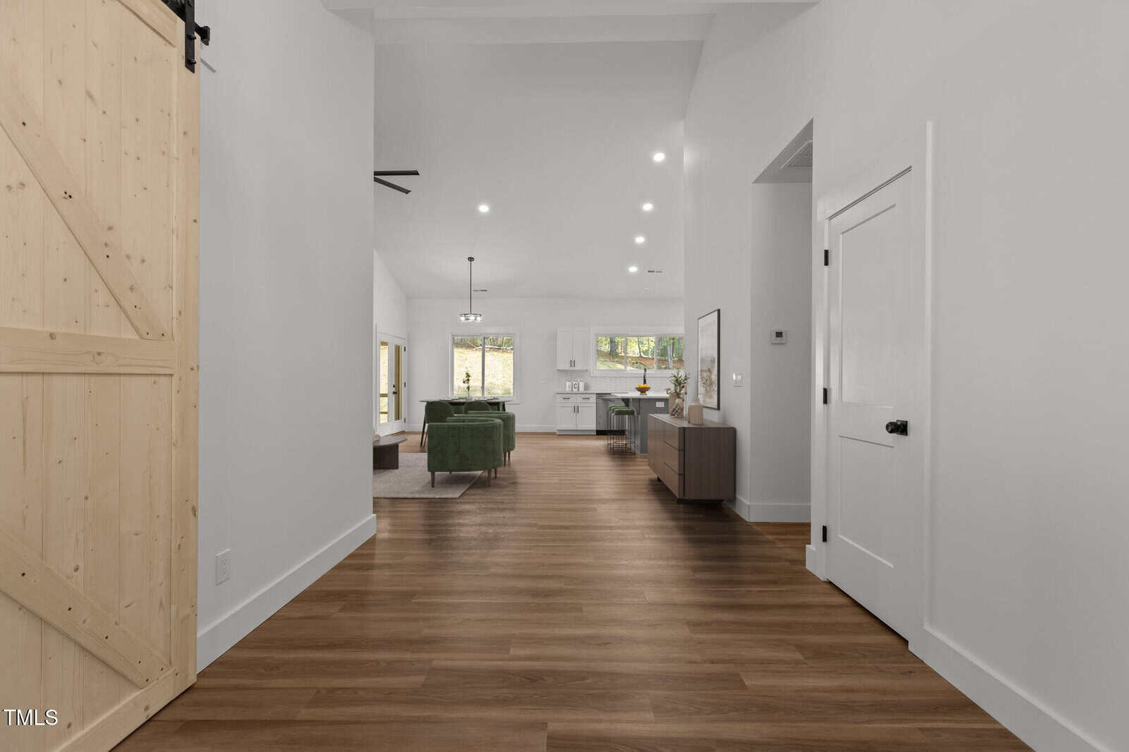 A hallway with light wood flooring leads to an open living space. The area features white walls, recessed lighting, a green sofa, kitchen appliances, and a wooden sliding barn door on the left.
