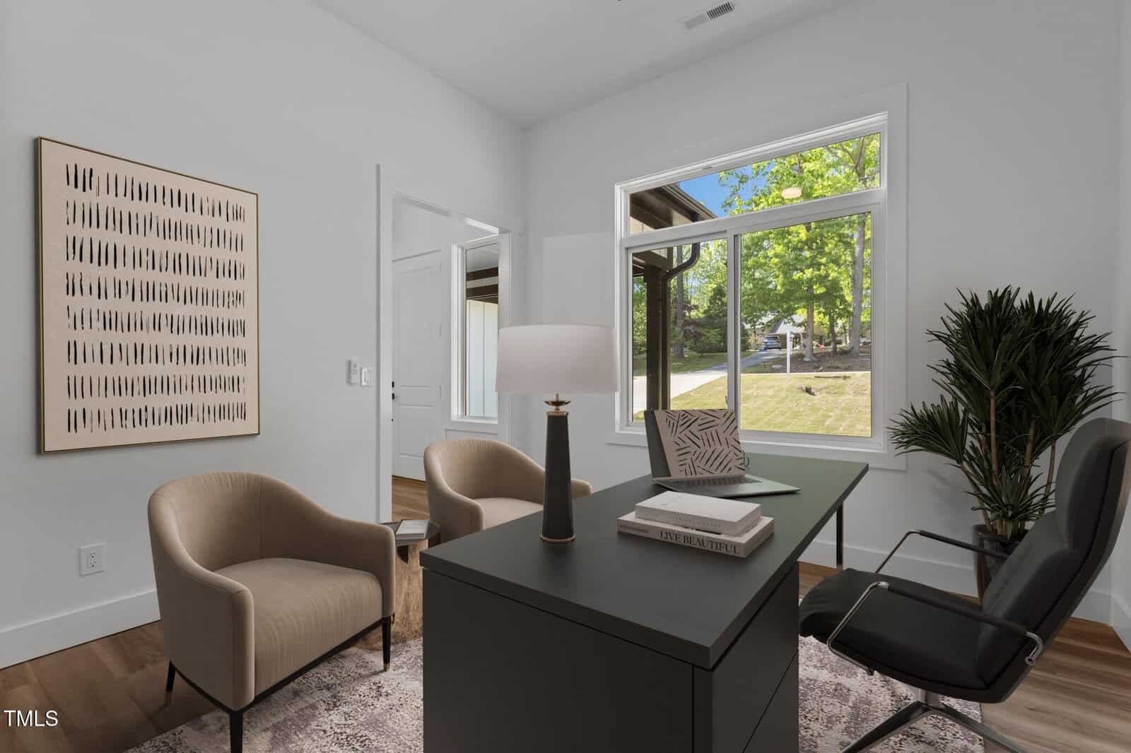 A modern office with a black desk, chairs, a lamp, and a plant. Large window lets in natural light and shows a green yard outside. Minimalist art is on the wall, and a door stands open in the background.