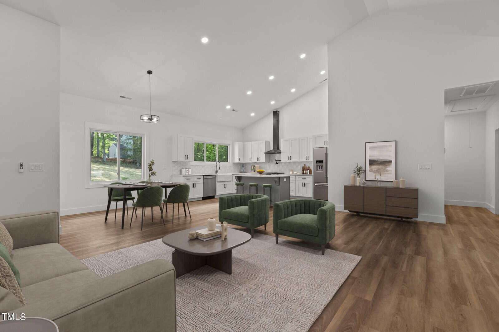 Modern open-concept living room and kitchen with white walls, wood flooring, green chairs, a beige sofa, and large windows. The kitchen features white cabinets, stainless steel appliances, and a central island.