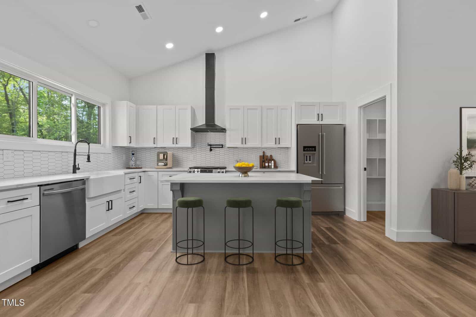 Modern kitchen with white cabinets, stainless steel appliances, island with three green barstools, and wood-style floors. A window above the sink lets in natural light. The space is bright and open with minimal decor.