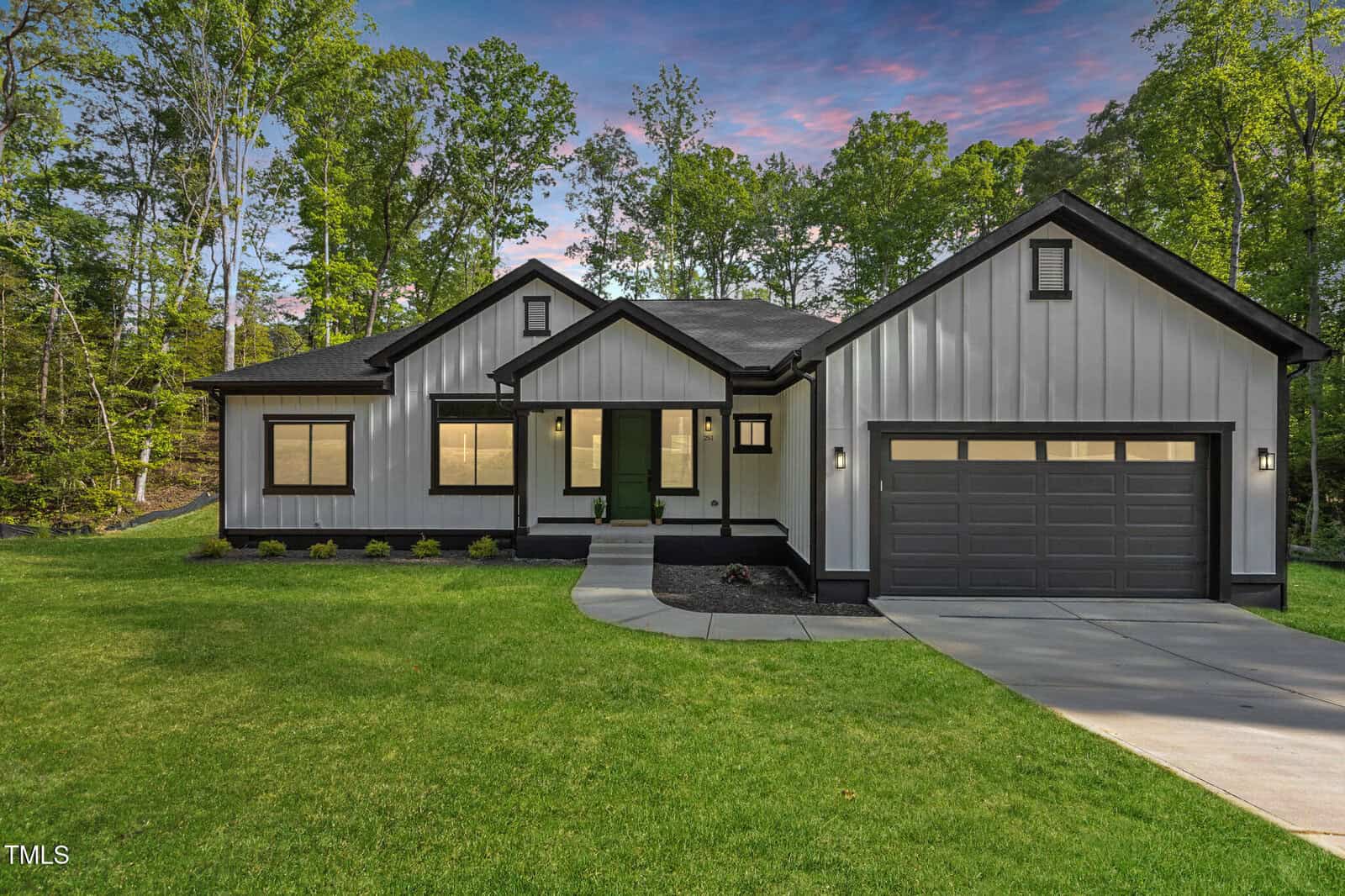 A modern single-story house with gray board-and-batten siding, black trim, a green front door, and a two-car garage, set on a manicured lawn bordered by trees under a colorful evening sky.