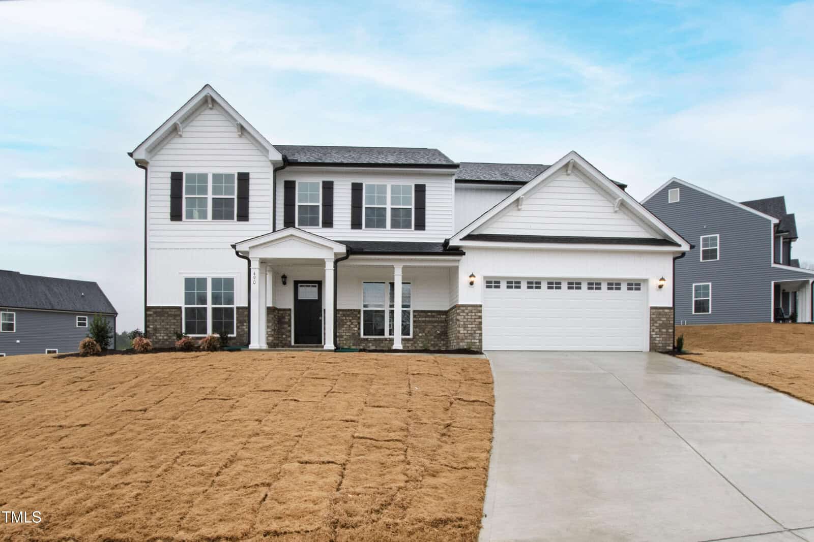Two-story white house with black trim and a covered front porch, stone accents, attached two-car garage, and a concrete driveway. The front yard has dry, brown grass and neighboring homes are visible in the background.