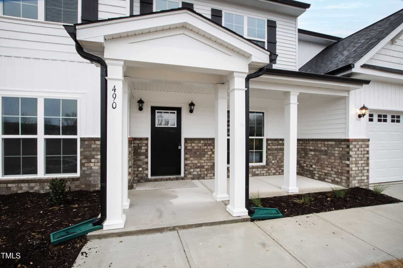 Front entrance of a modern two-story house with white siding, brick accents, black door, four white columns, and minimal landscaping along a concrete walkway. House number 490 is visible on a column.