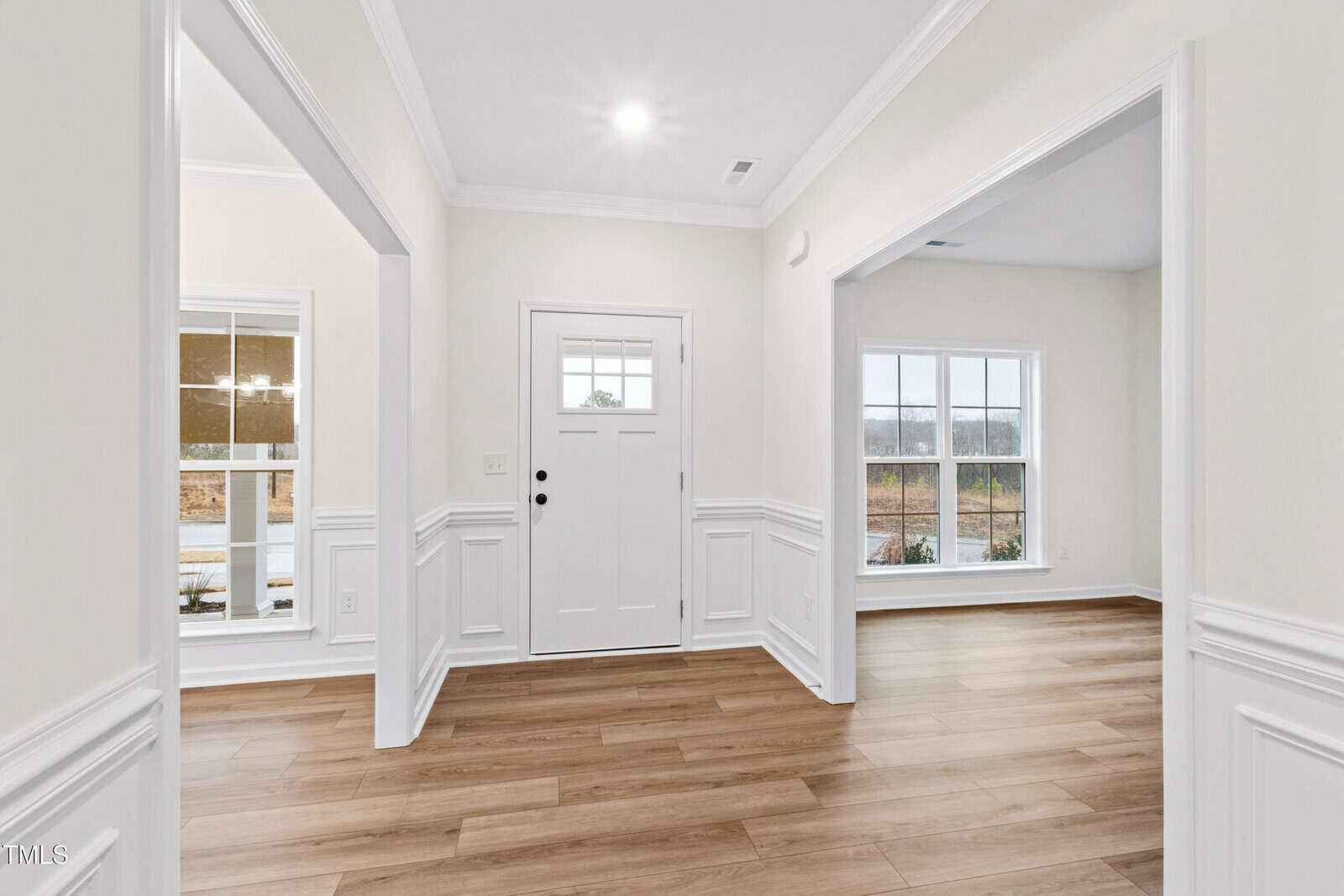 A bright entryway with light-colored walls, white trim, wood flooring, and large windows letting in natural light. The front door is centered, with two open rooms on either side.