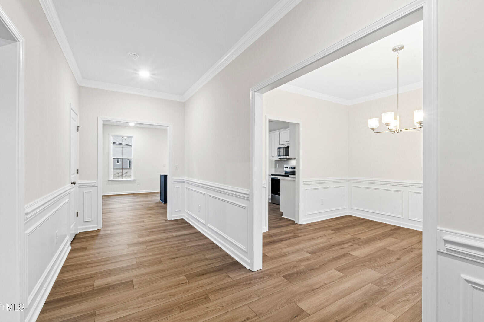 A bright hallway with light-colored walls, white trim, and wood-style flooring leads to a dining area with a chandelier and an open kitchen visible in the background.