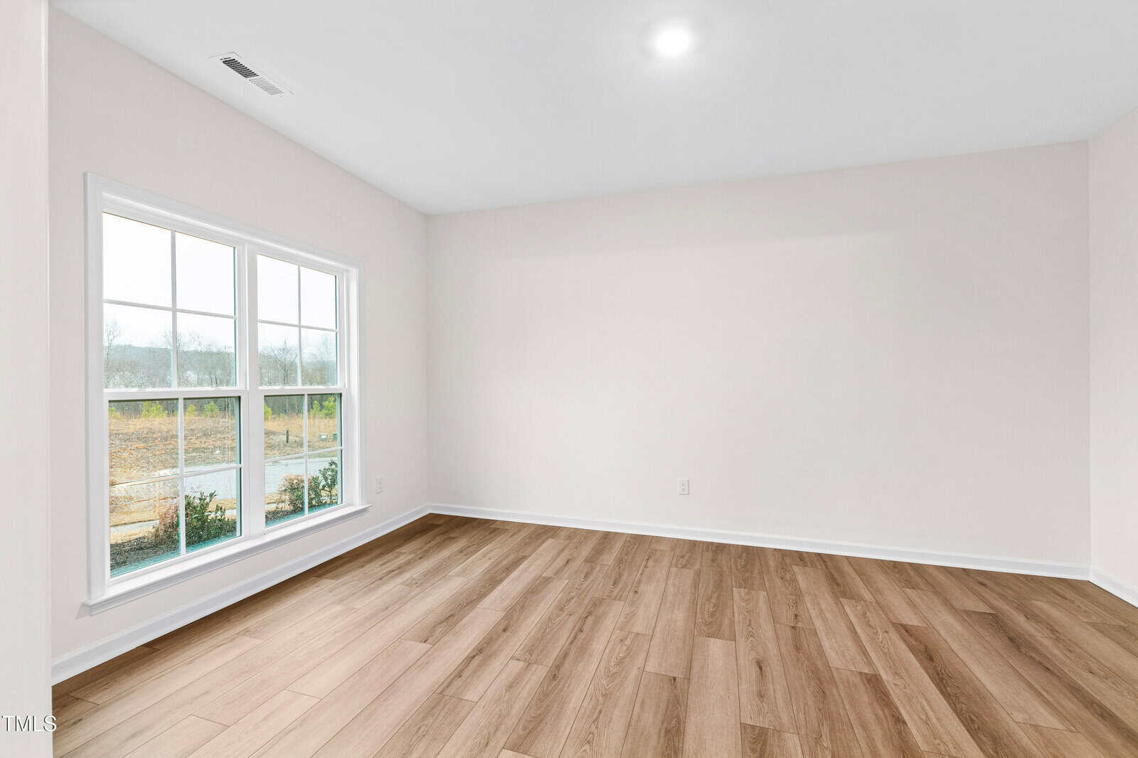 An empty room with light-colored walls, a large window letting in natural light, and wood-style flooring. The window shows an outdoor view of grass and trees. The ceiling has a single recessed light.