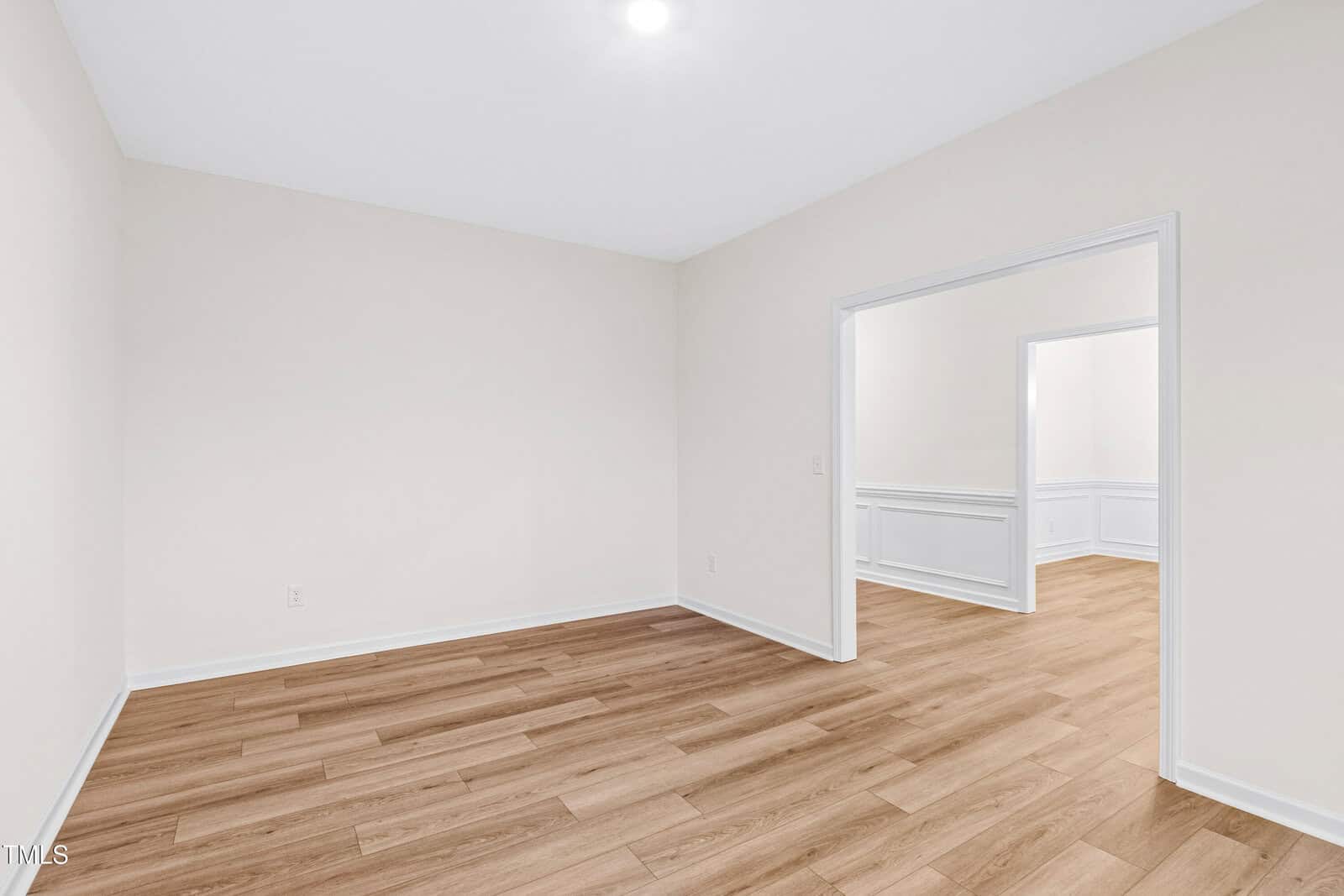 A bright room with light beige walls, wood-style flooring, and white trim. An open doorway leads to another similar room. Both rooms are empty and well-lit, with ceiling lights visible.