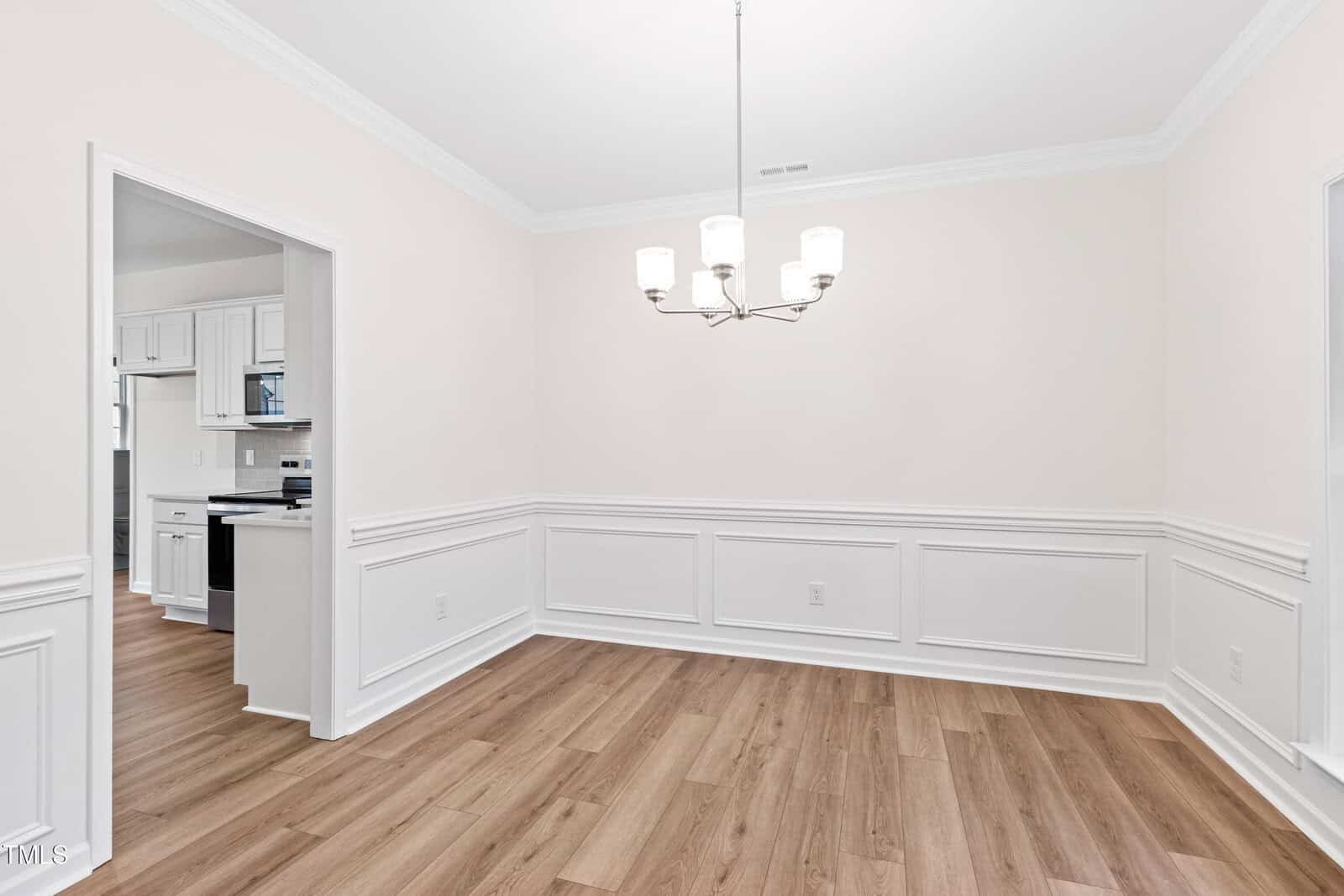 Bright dining room with light wood flooring, white wainscoting, and a five-light chandelier. The room opens into a kitchen with white cabinets and stainless steel appliances. Walls are painted off-white.