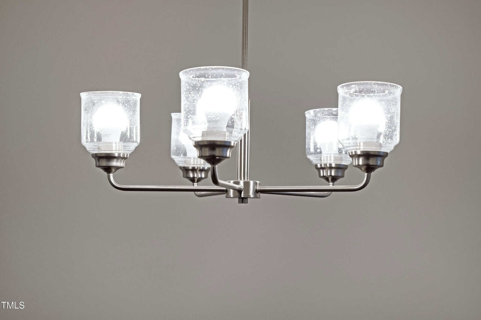 A modern chandelier with five lit bulbs, each covered by a clear glass shade, hangs from the ceiling against a plain gray background.
