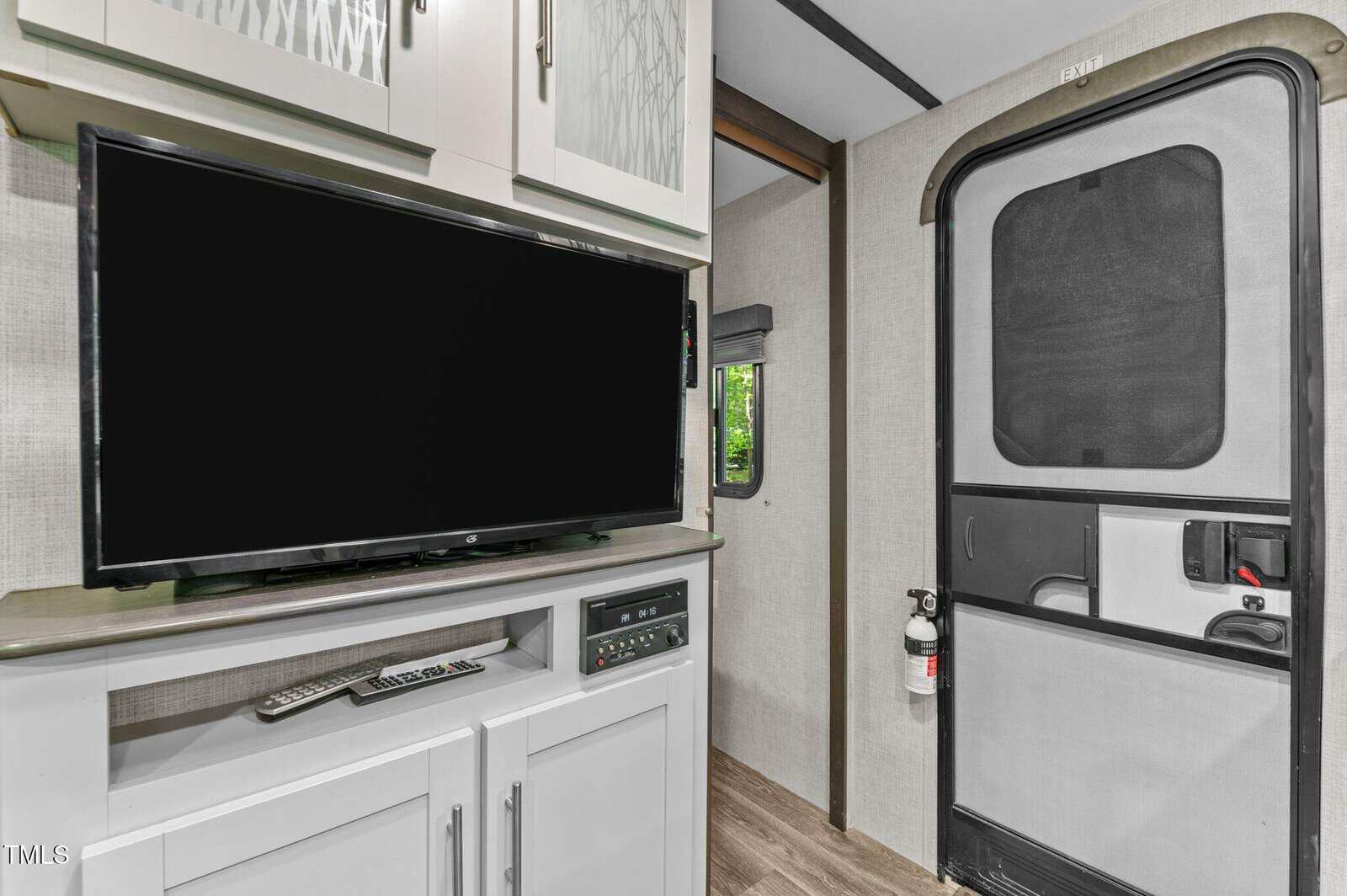 Flat-screen TV on a white cabinet, remote on the counter, electronic controls below, next to a door with a mesh screen and fire extinguisher; small window in the background showing green foliage.