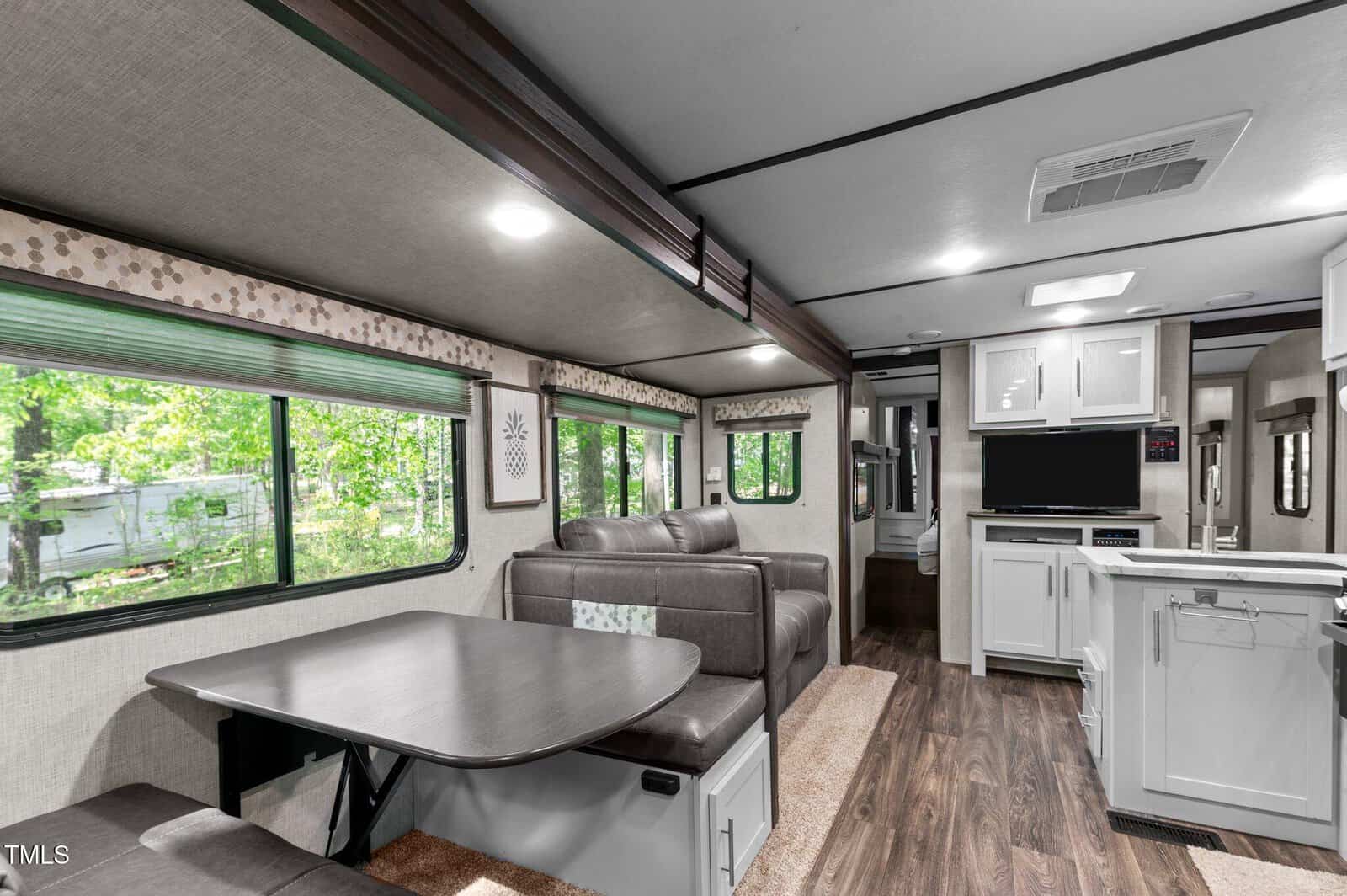 Interior of an RV featuring a dining area with a table and cushioned seating, a sofa, a kitchen with white cabinets, and windows with views of greenery outside. The floor is wood-style laminate.