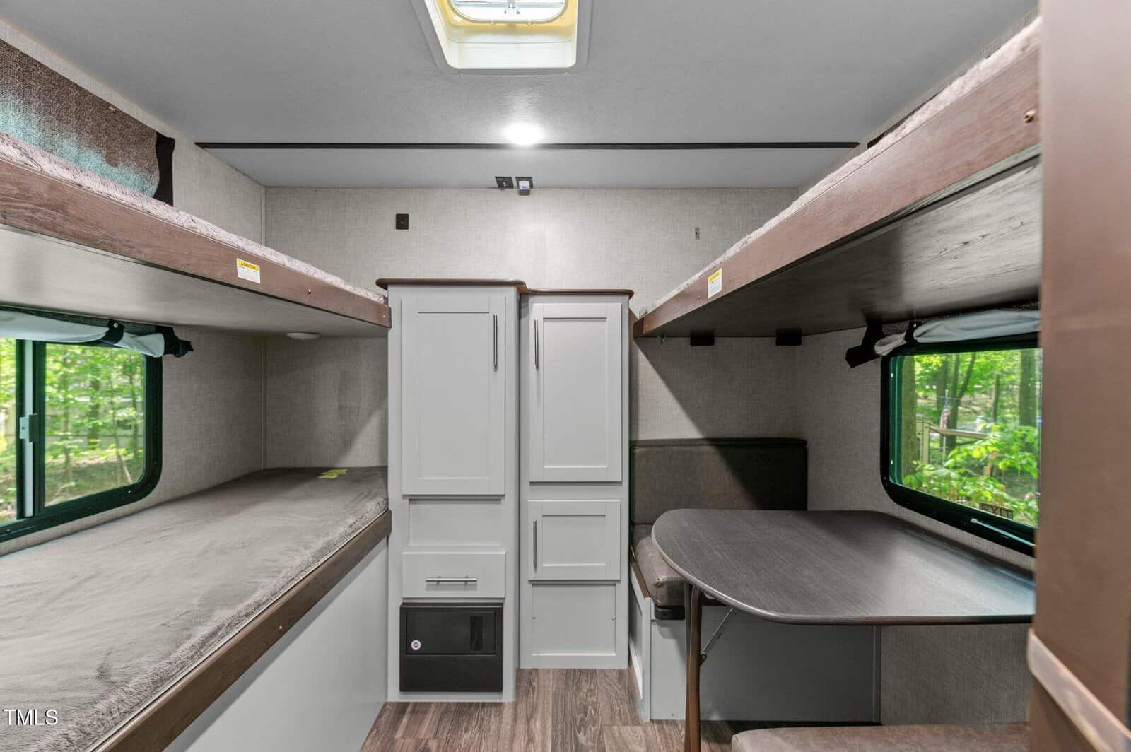 The interior of an RV features two bunk beds on either side, cabinets in the center, a small seating area with a table on the right, and windows with views of trees outside.