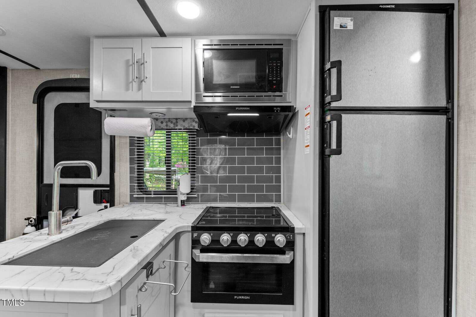 A compact kitchen features a marble-look countertop with a sink, a stainless steel faucet, a gas stove with oven, a microwave above, gray subway tile backsplash, upper cabinets, and a tall silver refrigerator.