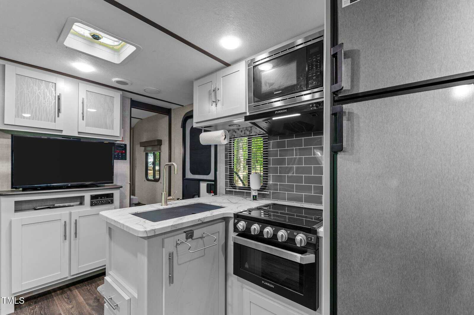 Modern RV kitchen with white cabinets, marble countertop, stainless steel appliances including fridge, oven, microwave, and a small sink. Gray subway tile backsplash, paper towel holder, TV, and window with blinds are visible.