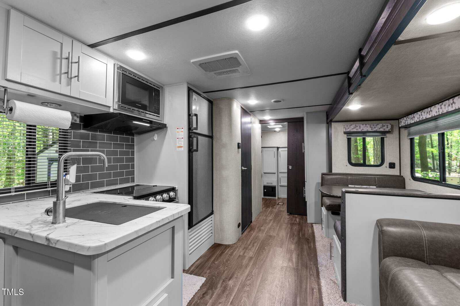 Interior of a modern RV with a white kitchen area, stainless steel sink, stovetop, microwave, gray tile backsplash, wood flooring, dining area, and windows with views of trees outside.