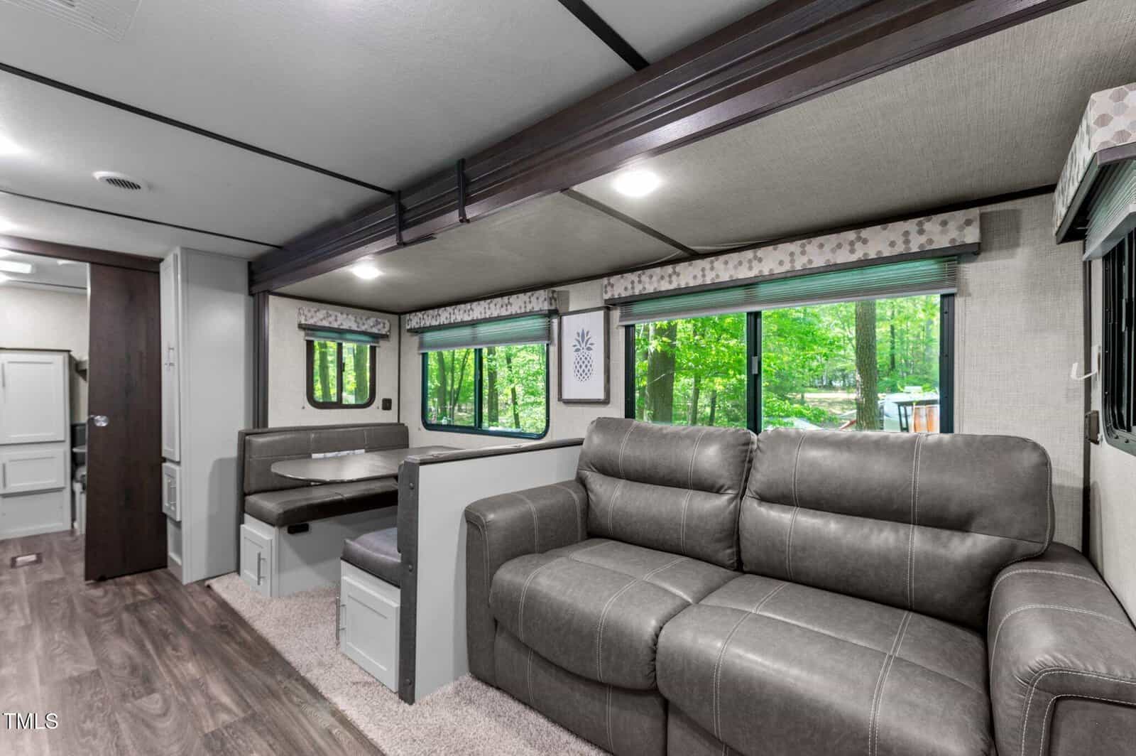 Interior of an RV with gray leather sofa, booth-style dining area, large windows showing green trees outside, and wood-style flooring. The space is well-lit and decorated in neutral tones.