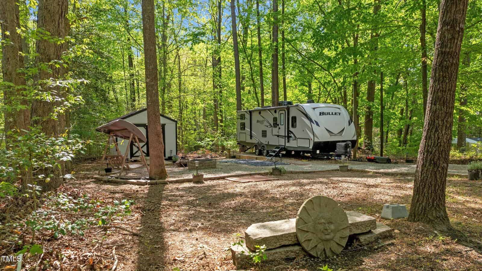 A white Bullet travel trailer is parked in a wooded campsite with a small shed, a covered swing, and a round stone sun sculpture in the foreground. Tall trees surround the area, and sunlight filters through the leaves.