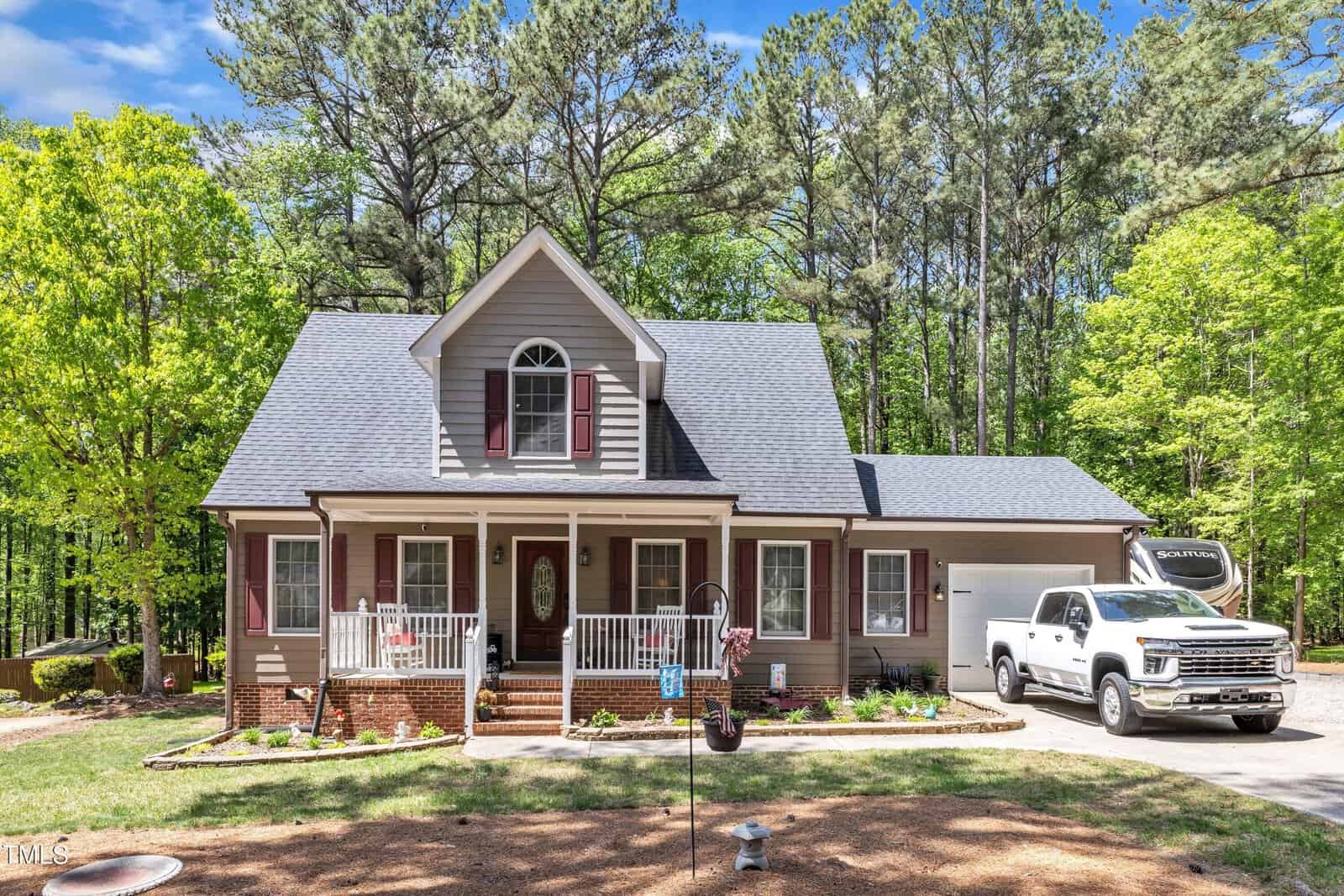 A beige house with a front porch, white railing, and red shutters is surrounded by trees. A white pickup truck with a trailer is parked in the driveway next to the house under a partly cloudy sky.