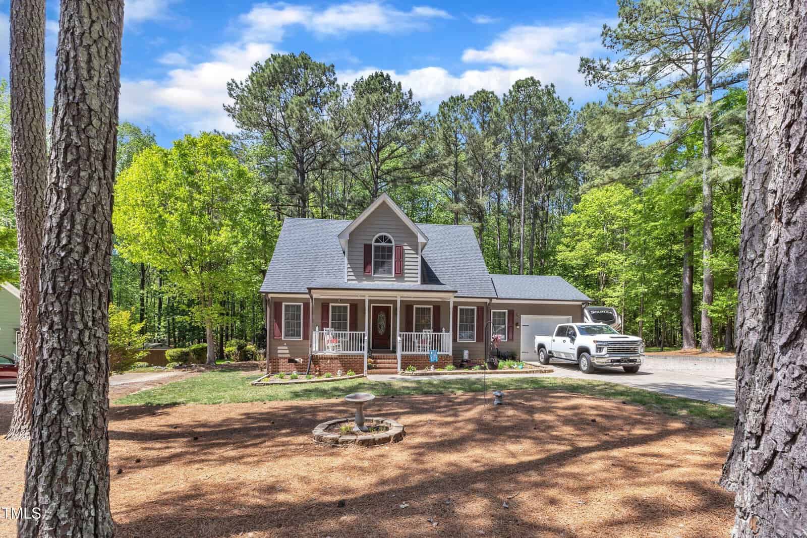 A two-story house with a porch and red shutters sits surrounded by tall trees. A white pickup truck is parked in the driveway, and a circular birdbath is in the front yard covered with pine straw.