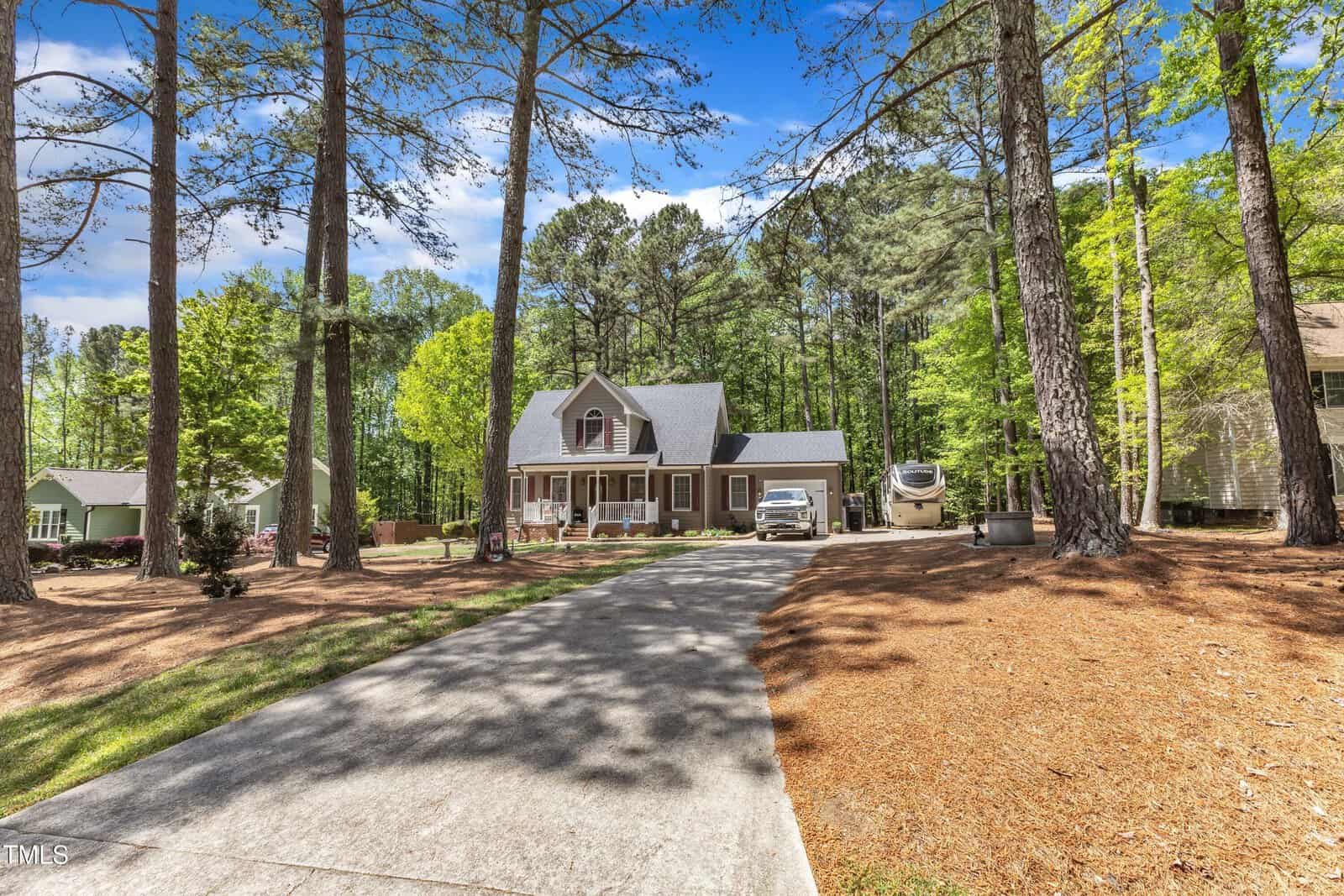 A two-story house with a front porch sits among tall trees. A concrete driveway leads up to the house, with a white vehicle and trailer parked nearby. The yard is landscaped with pine straw and surrounded by greenery.