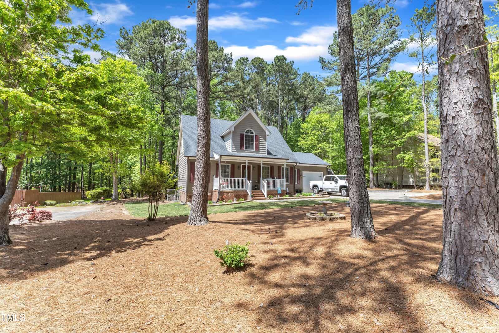 A two-story house with a covered front porch, gray siding, and gabled roof sits among tall trees with a pine straw-covered yard. A white pickup truck is parked in the driveway. The weather is clear and sunny.