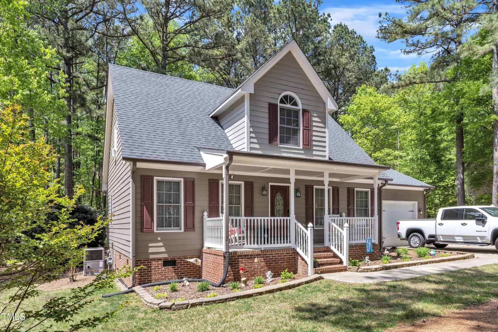 A two-story gray house with white trim, red shutters, and a covered front porch. There are steps leading to the porch, landscaped flower beds, and a white pickup truck parked in the driveway. Trees surround the house.