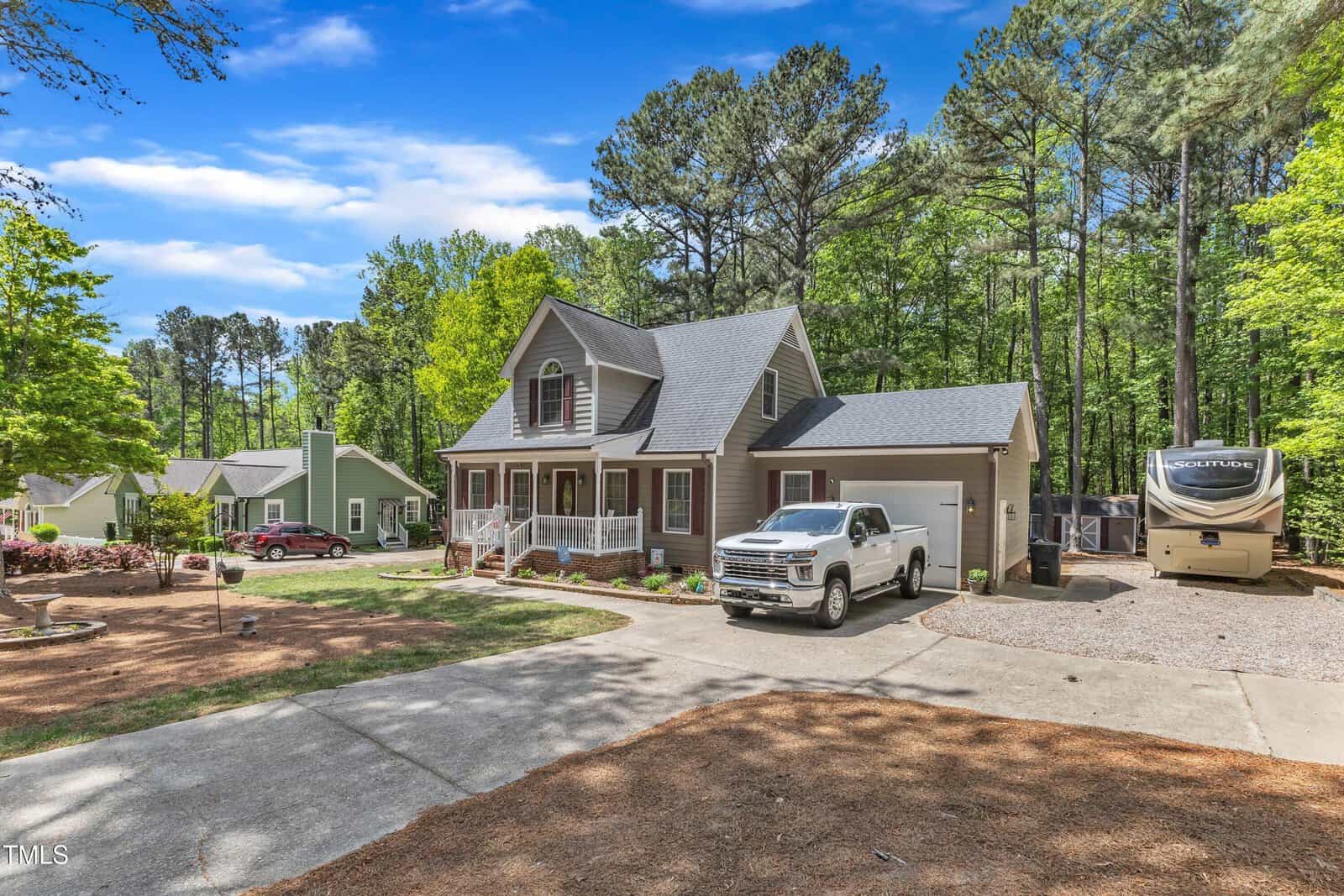 A two-story house with a front porch, a white pickup truck parked in the driveway, and an RV parked beside the house. The yard is landscaped, and tall trees surround the property under a clear blue sky.