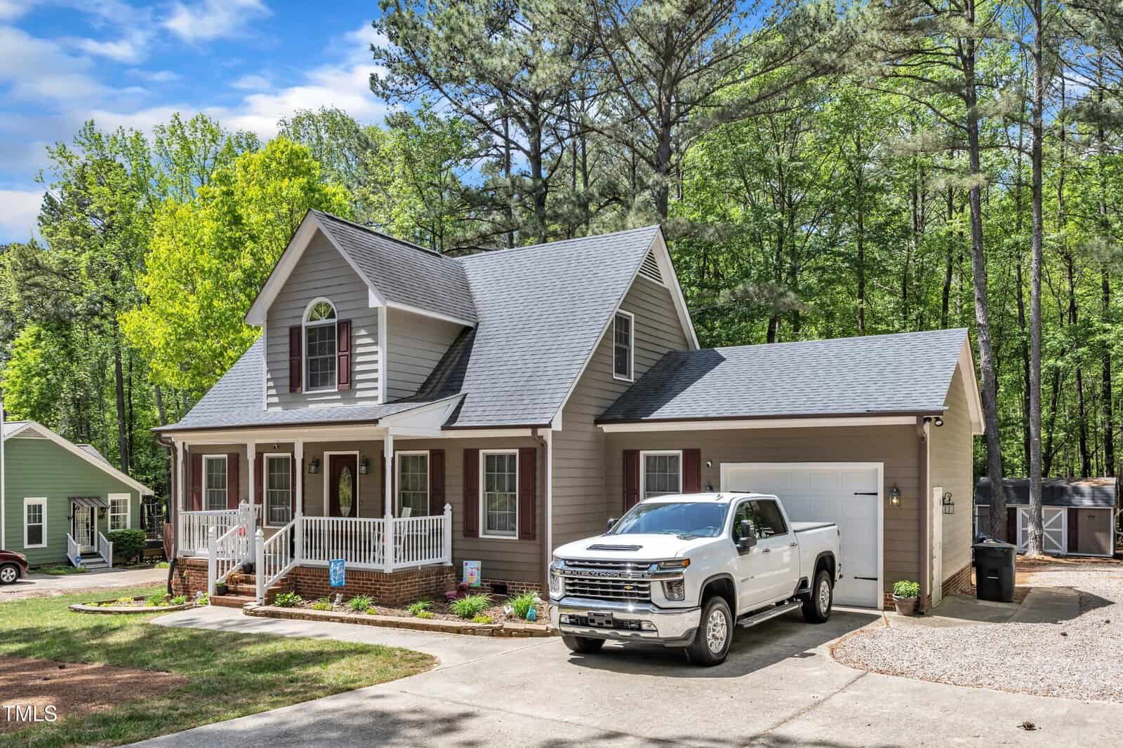 A two-story beige house with a front porch, red shutters, and a gable roof. A white pickup truck is parked in the driveway. The house is surrounded by trees and neighboring homes.