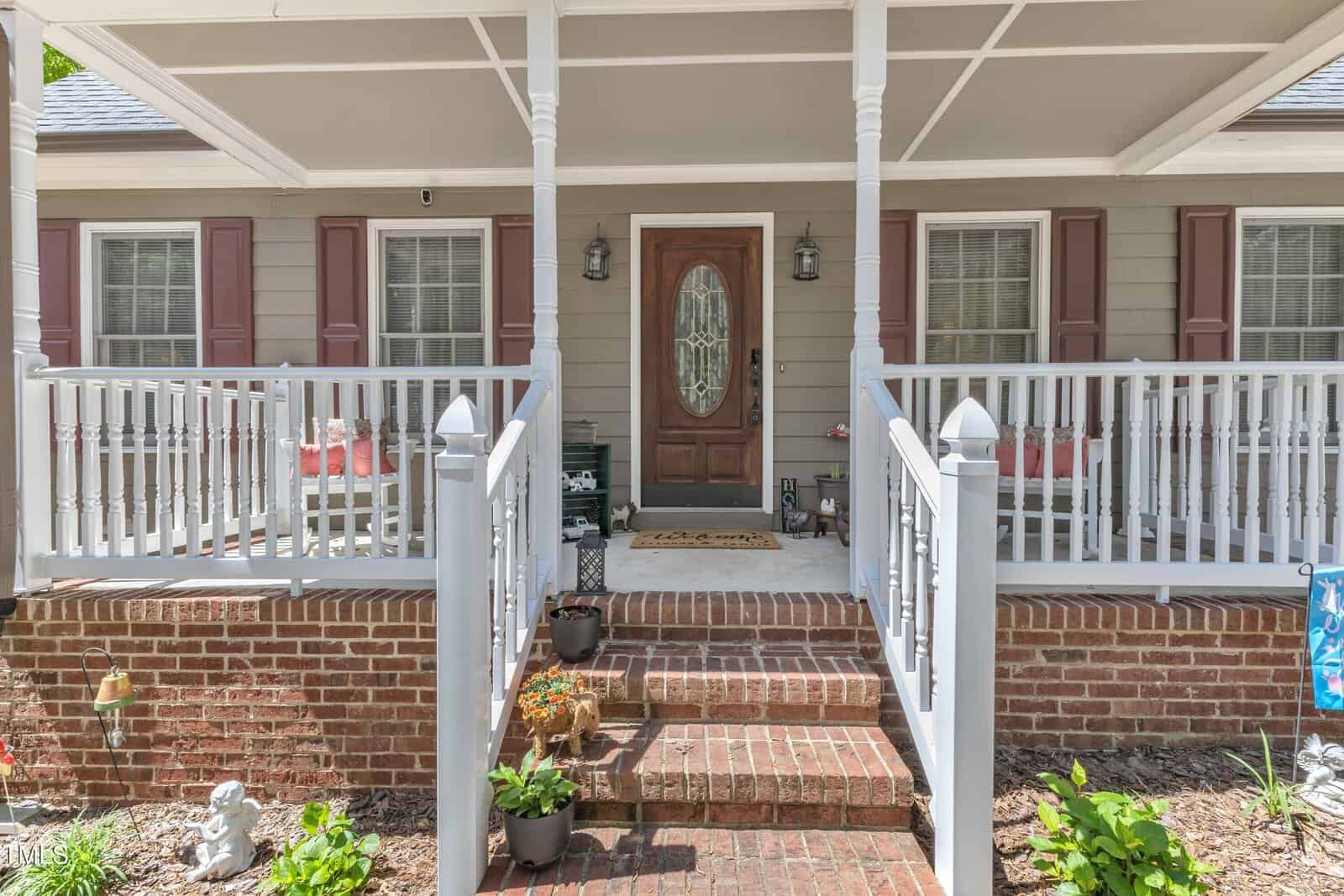 Front porch of a house with brick steps, white railings, gray siding, three windows with shutters, a brown wooden door with glass panel, potted plants, and decorative items placed near the entrance.