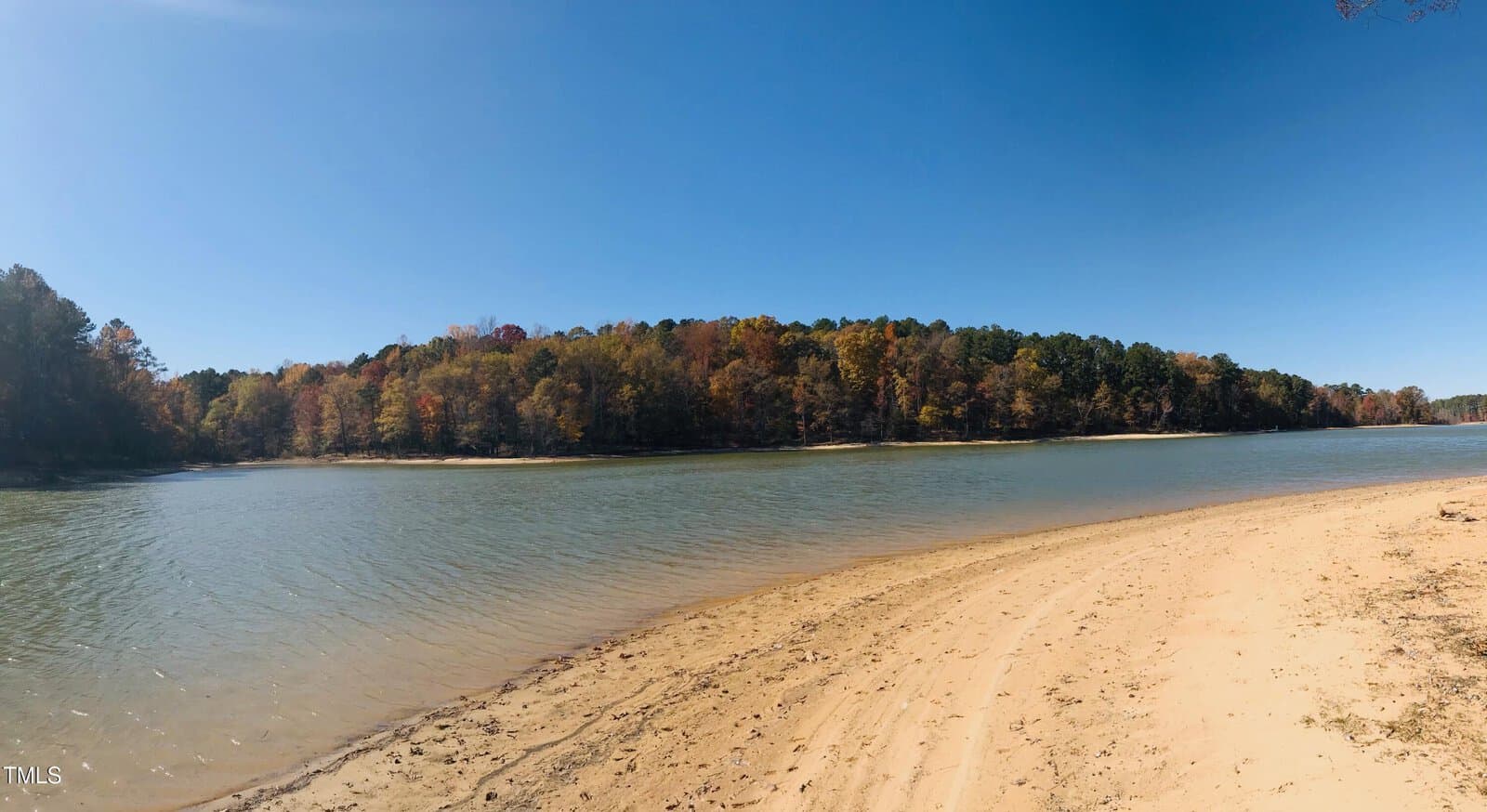 A calm lake with a sandy shoreline curves around the foreground. Dense trees with autumn foliage line the opposite shore under a clear blue sky.