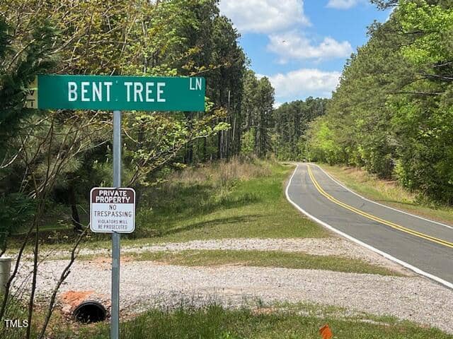 A green street sign labeled "BENT TREE LN" stands at a rural roadside with a "Private Property No Trespassing" sign below it. A two-lane road curves through a wooded area under a partly cloudy sky.