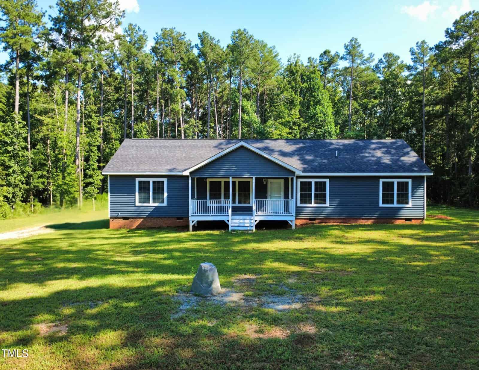 A single-story blue house with white trim sits on a grassy lawn at 1010 Pool Rock Road in Henderson, surrounded by tall trees. The house has a front porch with steps and railings, and multiple windows on the front facade.