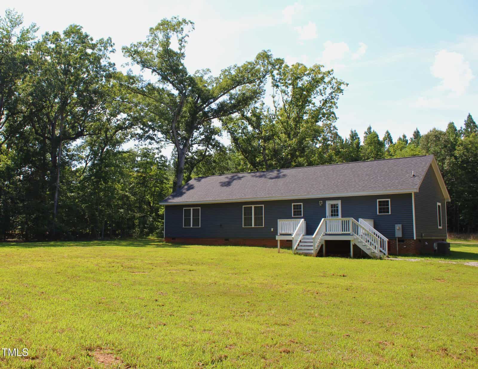 A single-story blue house with a dark roof sits on a large grassy lawn. White steps and railings lead to the front door. Trees surround the house, and the sky is partly cloudy.
