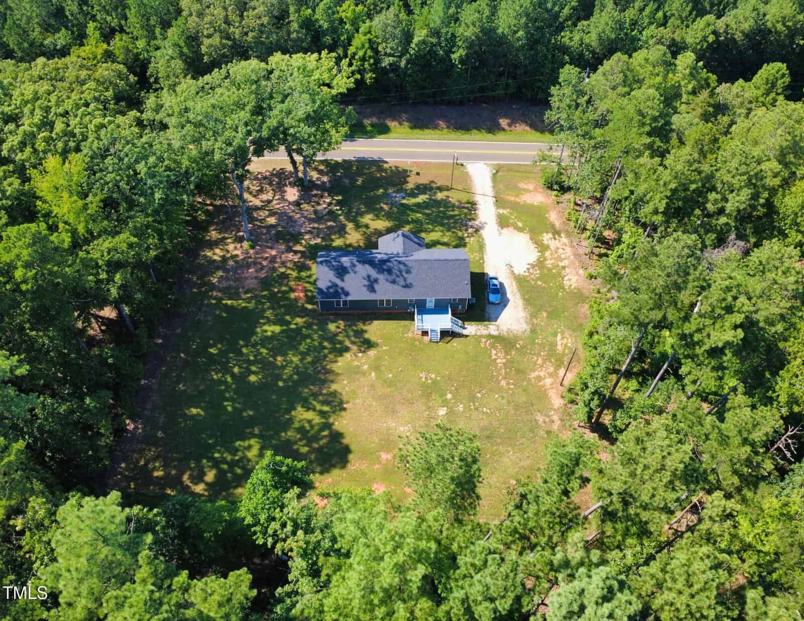 Aerial view of a single-story house with a dark roof, a driveway, and a small front porch, surrounded by trees and a grassy yard, with a road visible behind the property.