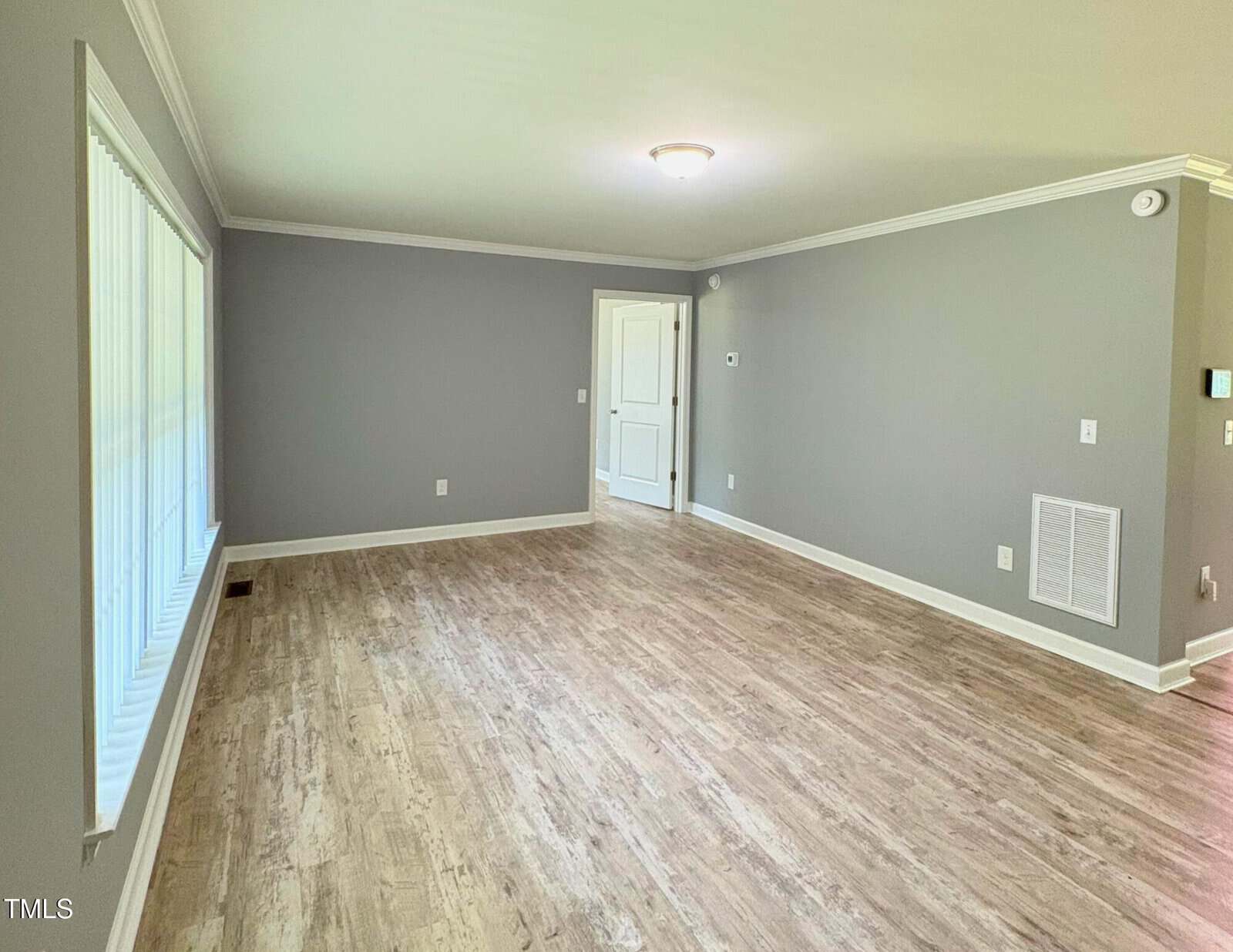 A bright, empty living room with light wood flooring, gray walls, white trim, and a ceiling light. Large windows line one wall, and a white door is visible at the far end of the room.