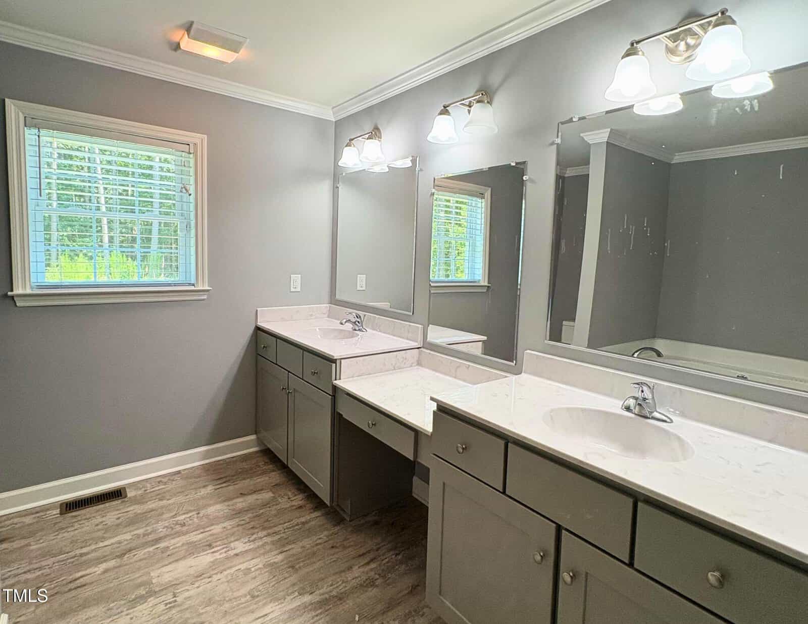 A bathroom with two sinks, gray cabinets, marble countertops, two mirrors, three light fixtures above the sinks, a window with blinds, and light-colored wood flooring.