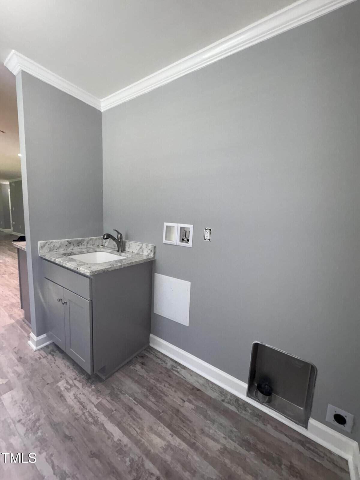 A laundry room with gray walls and flooring, a small gray cabinet with a sink and faucet, wall hookups for washer and dryer, and a metal dryer vent opening near the floor.