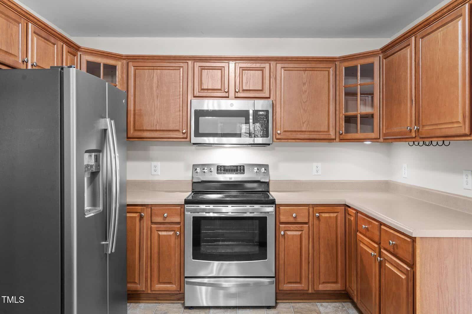 A kitchen with wooden cabinets, stainless steel refrigerator, electric stove, and microwave; beige countertops and tiled flooring are also visible.