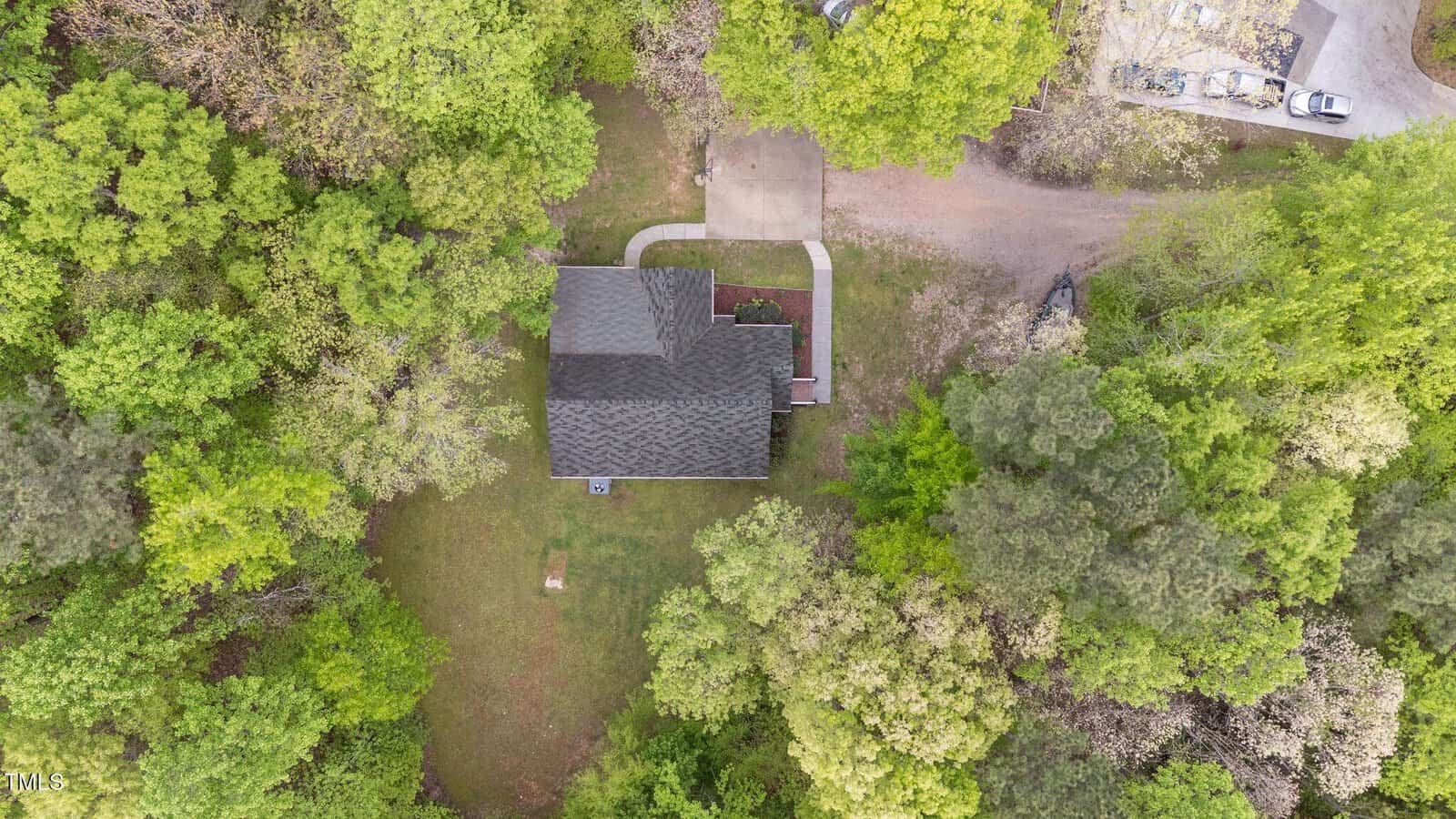 Aerial view of a house surrounded by green trees and lawn, with a driveway leading to a small parking area. A path curves from the driveway to the front entrance. A few vehicles are parked in the upper right corner.