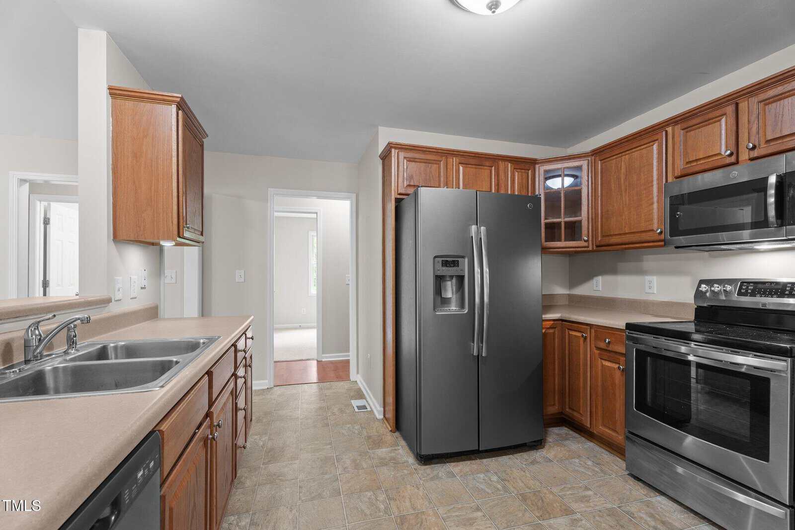 A kitchen with wood cabinets, stainless steel appliances including a double-door refrigerator, oven, microwave, and dishwasher, beige countertops, a double sink, and tiled flooring.