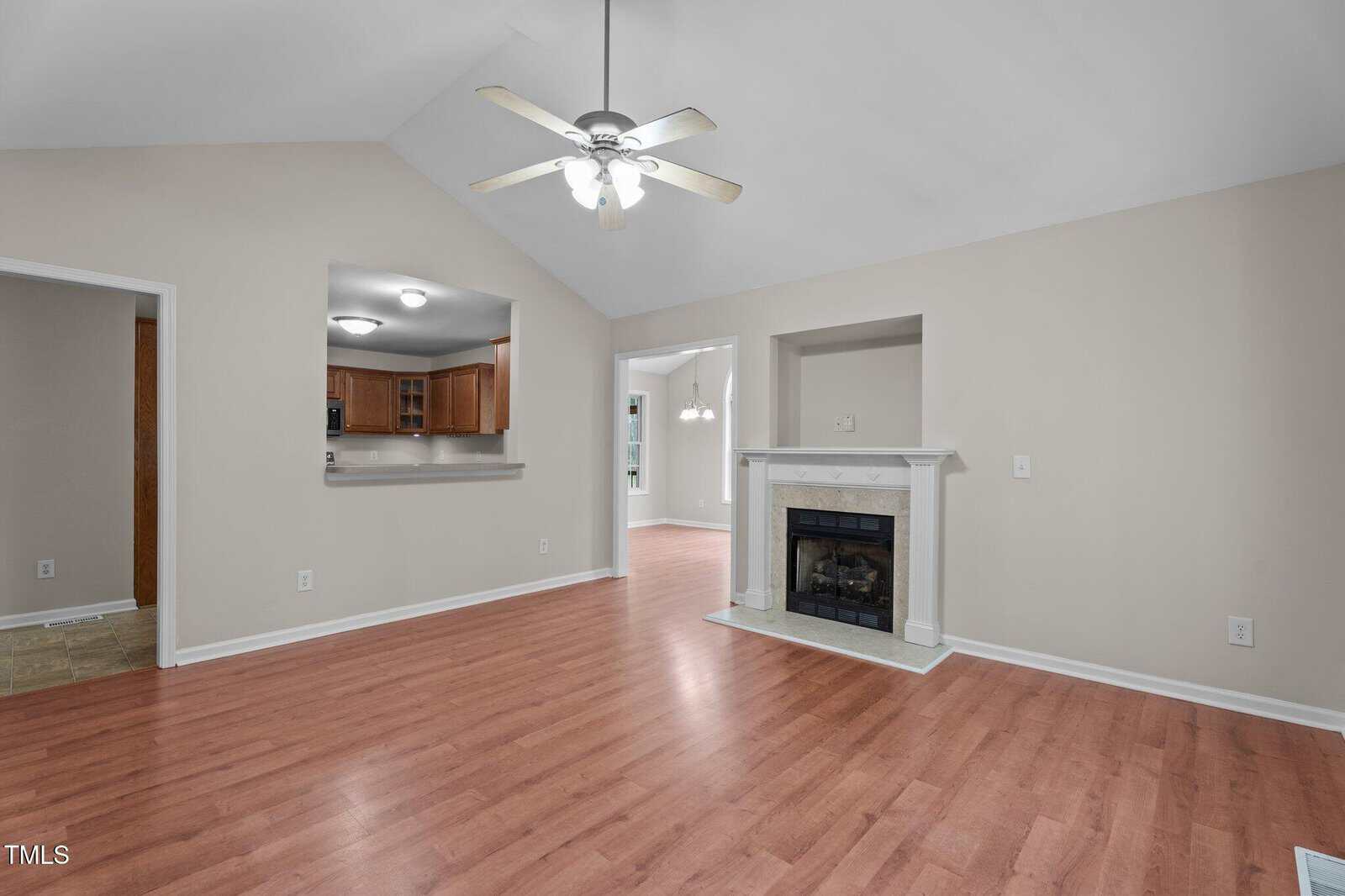 A living room with light-colored walls, wood-look flooring, a white ceiling fan, a fireplace with a white mantle, and open views into a kitchen and dining area through wide doorways.