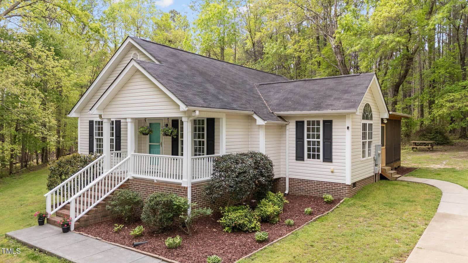 Single-story house with light-colored siding, dark shingle roof, white trim, and a small front porch with steps. The yard is landscaped with bushes and mulch, surrounded by trees and a concrete walkway.