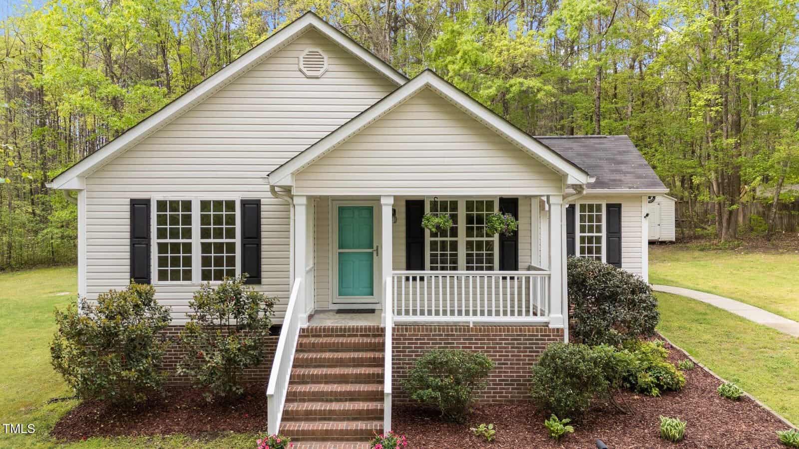 A single-story white house with black shutters, a teal front door, a small covered porch, brick steps, landscaped bushes, and a green lawn sits on 1106 Lake Ridge Drive in Creedmoor, surrounded by tall trees in the background.
