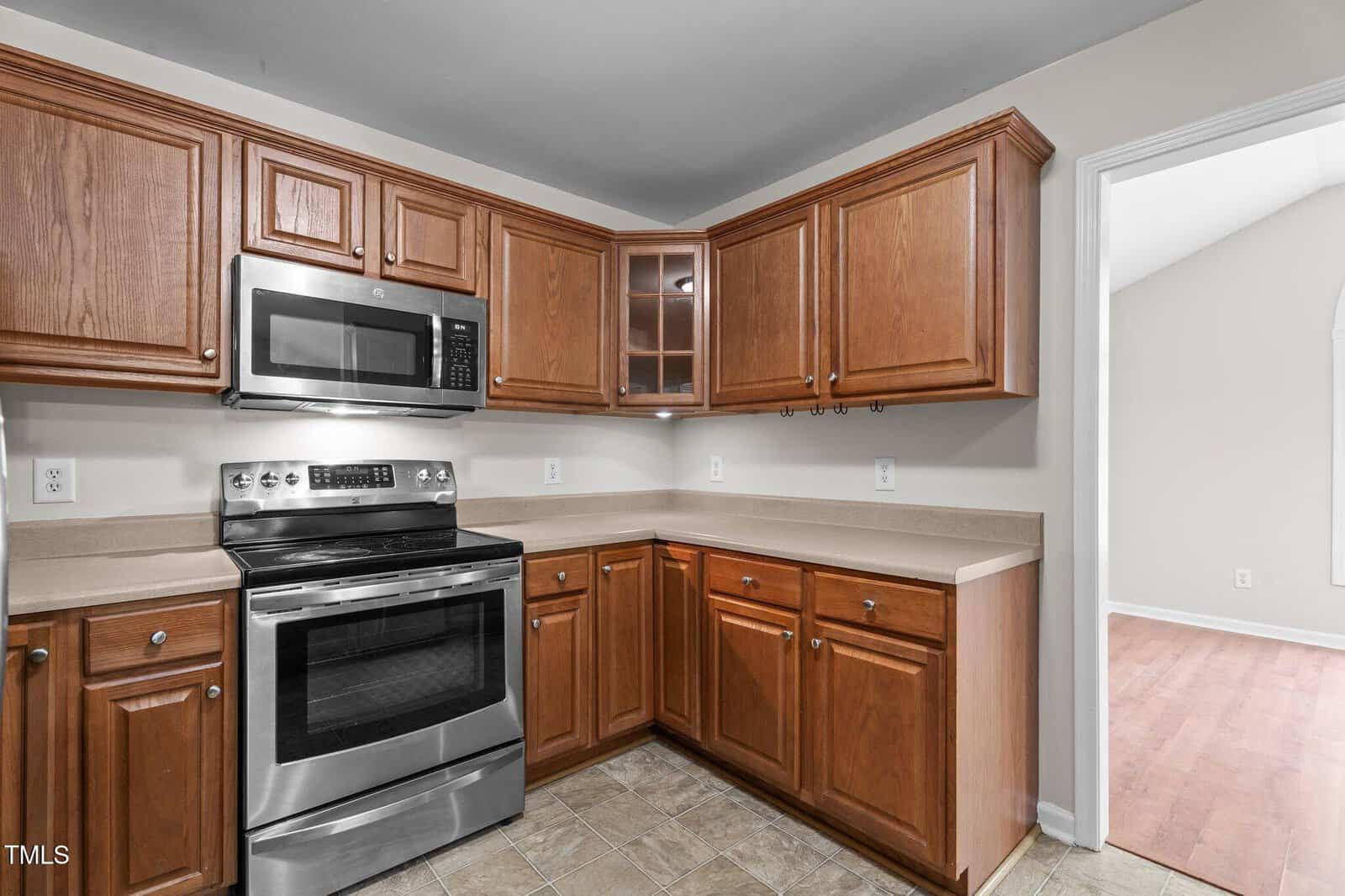 A kitchen with brown wooden cabinets, beige countertops, stainless steel oven and microwave, tiled floor, and an open doorway leading to a room with wood flooring.