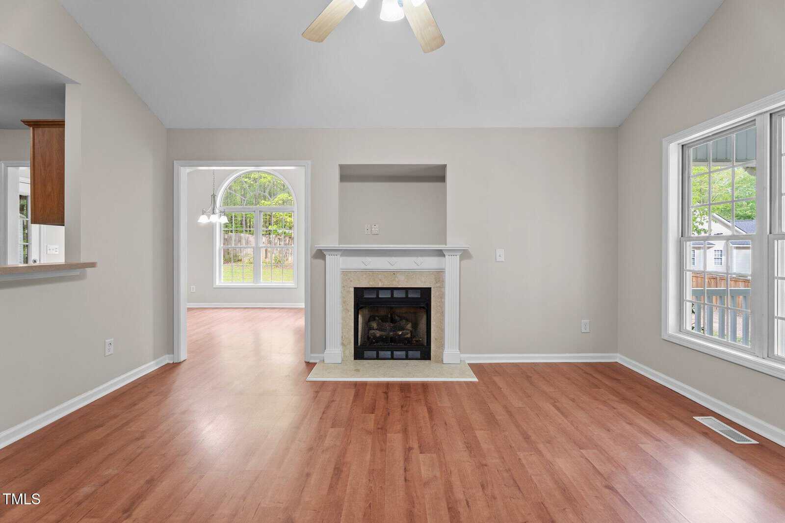 A living room with light beige walls, wood flooring, a white fireplace centered on one wall, and large windows on the right. An open entryway leads to a bright adjacent room with more windows.