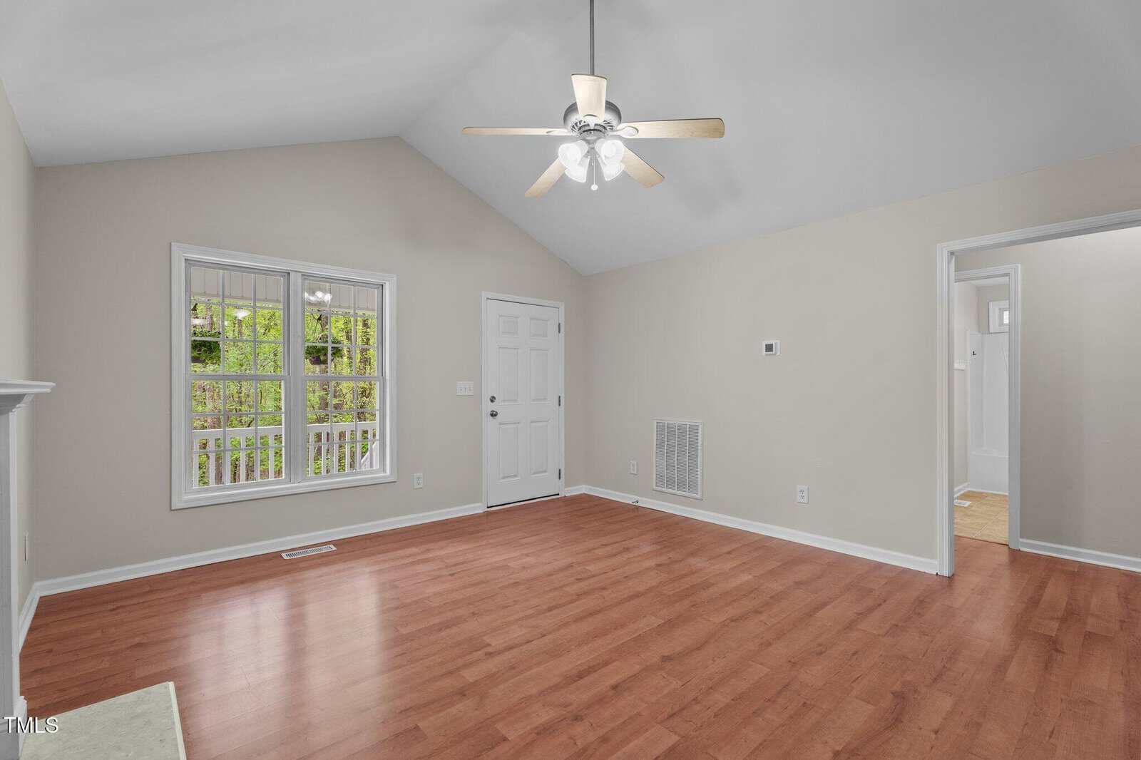 A bright, empty living room with light beige walls, wood flooring, a ceiling fan with lights, a white-framed window, and a white door. An open doorway leads to another room.
