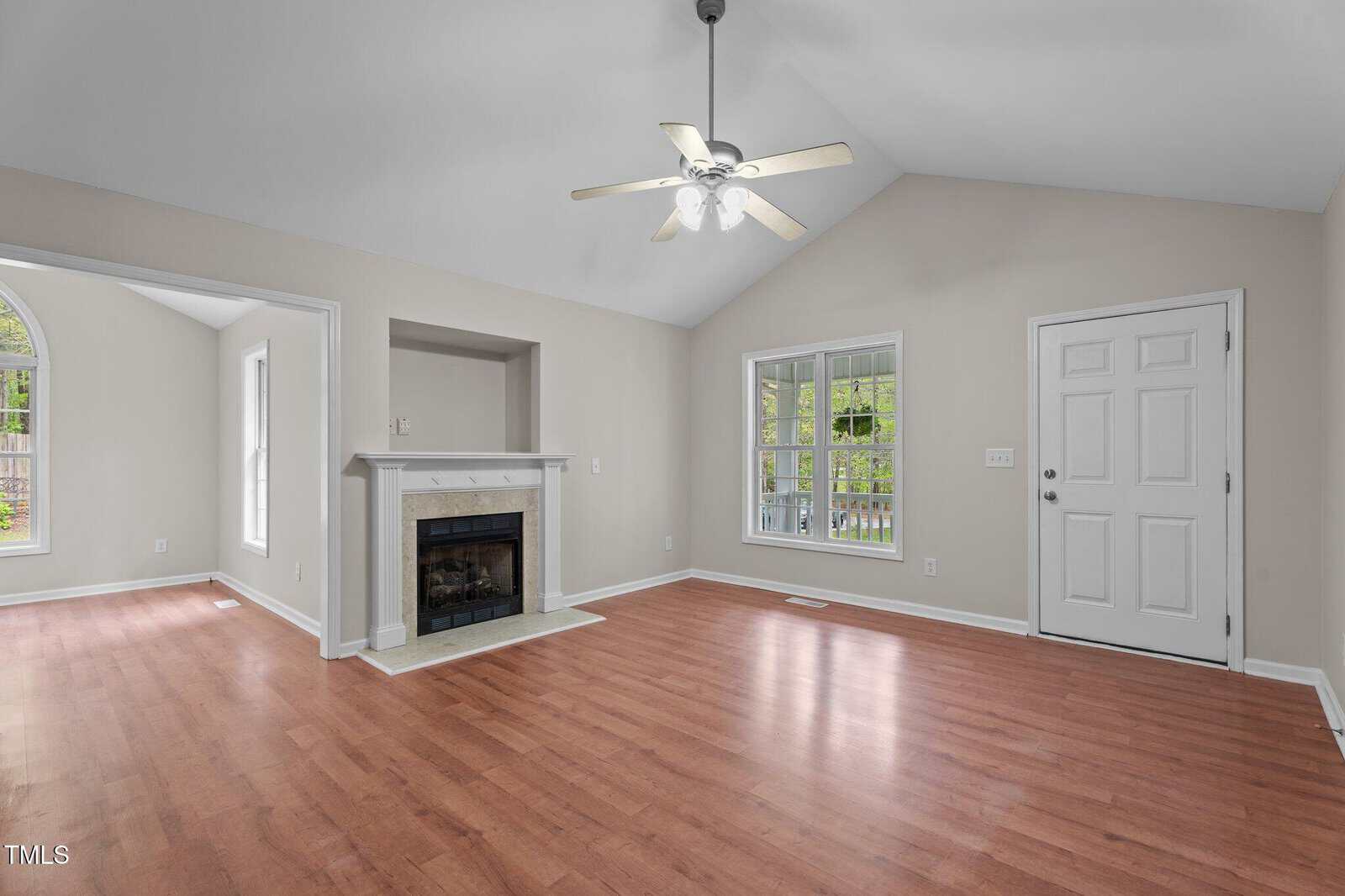 A living room with light beige walls, vaulted ceiling, ceiling fan with lights, white mantel fireplace, wood flooring, two large windows, and a white door; a bright adjacent room is visible through an open doorway.