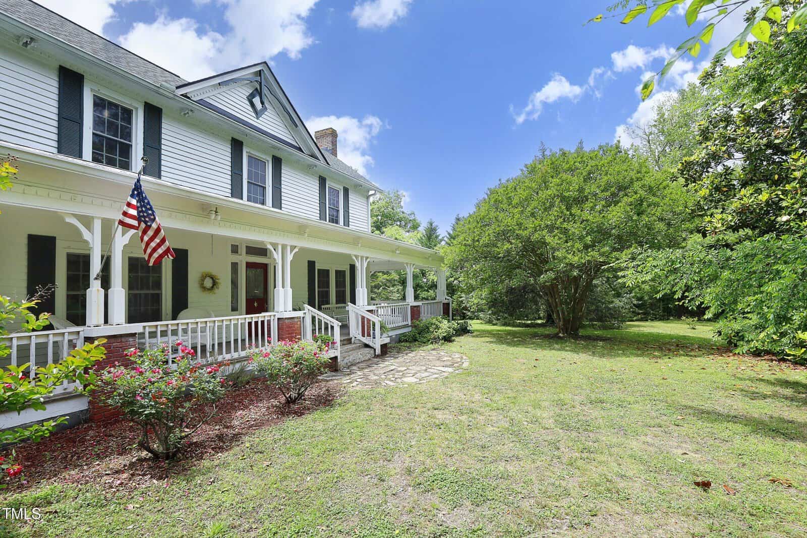 White two-story house with a large front porch, American flag, and brick accents; surrounded by green grass, shrubs, and trees under a blue sky with scattered clouds.