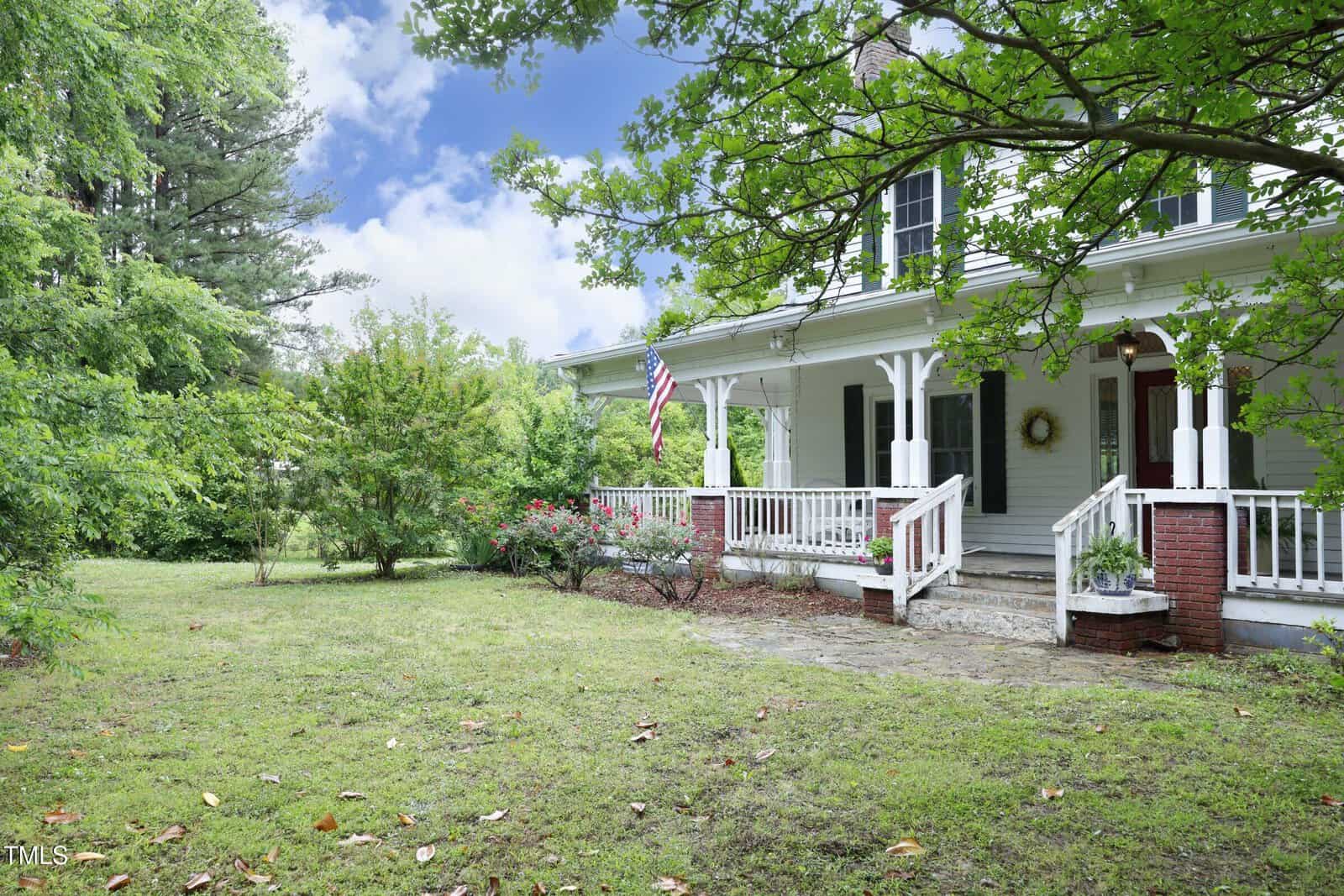 A white two-story house with a covered front porch, white railings, brick steps, and columns, surrounded by green trees and shrubs, with an American flag by the entrance.