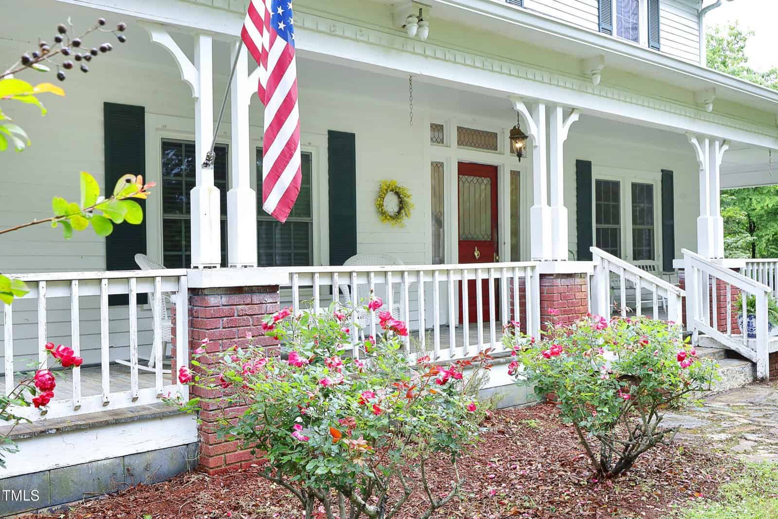 A white house with a large porch featuring white railings, red brick columns, and blooming rose bushes in front. An American flag hangs by the door, and a wreath decorates the front entrance.